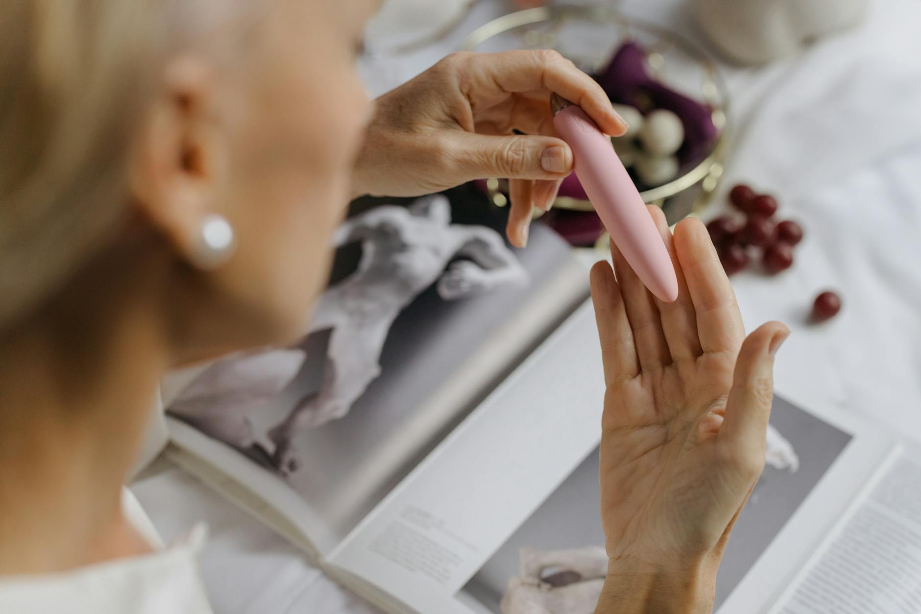 Close-up of hands holding a pink vibrator with art books nearby, symbolizing integrated pleasure into an intellectual, busy lifestyle.